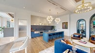 Located at Bennet at Bull Street in Columbia, SC, this thoughtfully designed single-floor space offers a generously proportioned open-plan layout that seamlessly integrates a fully equipped kitchen, multiple lounge areas, and distinct architectural features throughout.

The kitchen serves as a striking focal point, showcasing bold blue cabinetry paired with a large center island topped with a grey quartz surface and lined with black velvet bar stools. A dramatic dark marble-veined backsplash contrasts with shiplap-paneled upper walls, while globe chandelier pendants with brass fixtures illuminate the space from above. Stainless steel appliances complete the functional yet stylish setup.

The lounge areas are defined by curated furnishings, including deep tufted navy sofas, leather armchairs, and layered area rugs. A coffered ceiling with an ornate lattice detail and tiered chandelier anchors one seating zone, while floor-to-ceiling steel-framed windows flood the space with natural light and offer views to the exterior.

Distinctive arched niches adorned with botanical wallpaper and decorative lighting add an artistic, boutique-hotel quality to the space. Wide-plank hardwood flooring runs throughout, tying each zone together cohesively and contributing to the space's refined, upscale ambiance.