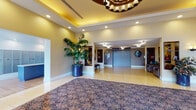 This well-appointed building lobby offers a polished and inviting first impression through thoughtful architectural design and curated décor. The entrance foyer is framed by floor-to-ceiling glass walls and doors, flooding the space with natural light and offering views of the surrounding greenery outside. Underfoot, large-format terrazzo tiles extend throughout, accented by a richly patterned area rug that anchors the central gathering space. A coffered ceiling with warm yellow recessed lighting adds depth and a sense of grandeur to the main entry area, complemented by a decorative chandelier overhead. A staffed reception desk with a rich wood-toned facade serves as the functional hub of the lobby, positioned to greet all who enter. Built-in display niches along the walls feature decorative sculptures, artwork, and colorful paintings that add artistic character throughout. Lush tropical potted plants, including a tall palm, bring life and warmth to the space. A seating alcove with upholstered armchairs and a side table provides a comfortable waiting area adjacent to the corridor leading to elevators. The elevator vestibule is cleanly finished and well-lit, offering efficient access to upper floors. A long, carpeted hallway extends from the lobby, lined with evenly spaced doors. Overall, the lobby balances functionality with an elegant, welcoming ambiance.