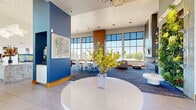 This contemporary clubhouse in Denver, Colorado offers a generously proportioned single-floor gathering space designed for community interaction and relaxation. The open-plan layout is anchored by soaring ceilings and expansive floor-to-ceiling windows that flood the interior with natural light and provide sweeping views of the surrounding outdoor areas.

The main social space is divided into several distinct functional zones that flow seamlessly into one another. A lounge area features plush, modern seating in cool blue-gray tones set atop richly patterned carpet, accompanied by a sleek fireplace surround clad in wood-toned paneling and a wall-mounted television. A billiards table adds a recreational focal point at the heart of the space. Separate seating nooks, including a cozy sofa arrangement near natural-light-filled windows, offer quieter retreats within the larger room.

The full kitchen is a standout feature, boasting granite countertops, a large central island with bar seating, crisp white cabinetry, a decorative patterned tile backsplash, stainless steel appliances, and tall windows that frame lush treetop views. A dining area with modern table and chairs sits adjacent. A vibrant living green wall adds a striking biophilic design element. Throughout, the palette of deep blues, warm wood accents, and bright whites creates an inviting, upscale ambiance.