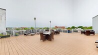 This spacious rooftop deck in Baltimore, MD offers an elevated outdoor retreat with sweeping panoramic views of the city skyline and surrounding neighborhoods. The deck is finished with warm-toned composite wood planking that spans a generous open area, providing ample room for both relaxation and social gatherings.

Multiple seating arrangements are thoughtfully distributed across the deck, including wicker dining sets with glass-top tables ideal for outdoor meals, as well as additional lounge groupings for casual conversation. A row of adjustable sun loungers lines one side of the deck, perfect for enjoying open-air leisure. Classic lamp post fixtures are positioned throughout, adding a charming architectural detail and evening illumination.

The deck is enclosed by a clean white railing that frames unobstructed views of Baltimore's urban landscape, including notable city landmarks visible on the horizon. A distinctive vintage water tower bearing the "Sail Cloth" insignia serves as a unique historic focal point on the rooftop. Access to the interior is provided through a set of French doors flanked by wall-mounted lanterns along the building's exterior facade.

Potted plants and floral arrangements add touches of greenery and color throughout the space, enhancing the welcoming atmosphere of this well-appointed rooftop amenity.