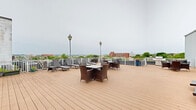 This spacious rooftop deck in Baltimore, MD offers an elevated outdoor retreat with sweeping panoramic views of the city skyline and surrounding neighborhoods. The deck is finished with warm-toned composite wood planking that spans a generous open area, providing ample room for both relaxation and social gatherings.

Multiple seating arrangements are thoughtfully distributed across the deck, including wicker dining sets with glass-top tables ideal for outdoor meals, as well as additional lounge groupings for casual conversation. A row of adjustable sun loungers lines one side of the deck, perfect for enjoying open-air leisure. Classic lamp post fixtures are positioned throughout, adding a charming architectural detail and evening illumination.

The deck is enclosed by a clean white railing that frames unobstructed views of Baltimore's urban landscape, including notable city landmarks visible on the horizon. A distinctive vintage water tower bearing the "Sail Cloth" insignia serves as a unique historic focal point on the rooftop. Access to the interior is provided through a set of French doors flanked by wall-mounted lanterns along the building's exterior facade.

Potted plants and floral arrangements add touches of greenery and color throughout the space, enhancing the welcoming atmosphere of this well-appointed rooftop amenity.