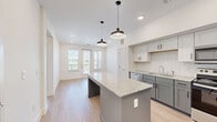 Located on N Grand Ave in Gainesville, Texas, this single-story, one-bedroom, one-bathroom unit offers a thoughtfully designed layout that maximizes its compact footprint. The open-concept kitchen is a standout feature, showcasing two-tone cabinetry in soft gray and white, stainless steel appliances including a refrigerator with ice maker, electric range, and microwave. A herringbone-patterned tile backsplash and granite countertops add a refined touch, while a central island with pendant lighting provides additional prep space and serves as a natural focal point. Light wood-look flooring flows throughout the main living areas, enhancing the bright, airy atmosphere created by recessed lighting and white walls. The bedroom is carpeted for comfort, fitted with a ceiling fan, and benefits from a window that invites natural light and outdoor views. A dedicated closet provides practical storage. The bathroom is well-equipped with a dark-cabineted vanity topped with granite, a large mirror, and a glass-enclosed walk-in shower with tile surround. A connecting hallway links all rooms efficiently, contributing to a clean, functional flow throughout the unit.