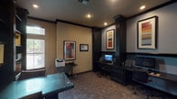 This business center is a single-level workspace designed to accommodate a variety of professional needs. The interior is defined by sophisticated dark wood architectural details, including fluted columns, crown molding, and dark wood trim that frame the space throughout. The flooring consists of a patterned carpet that adds texture underfoot.

The workstation area features two built-in computer desks finished in dark wood, each equipped with a monitor and ergonomic chair, positioned along one wall beneath a pair of boldly colored framed art pieces. A printer station on a slim metal stand offers shared printing access nearby.

A central conference area is anchored by a substantial glass-topped table with dark column-style legs, surrounded by task chairs, making it well-suited for meetings or collaborative work. Flanking built-in bookshelves with dark cabinetry below display decorative objects, adding a curated, professional aesthetic.

An entry corridor with glass-paneled double doors connects the business center to adjacent common areas, with natural light visible beyond. A posted business center policy board near the entrance provides clear usage information. Recessed lighting illuminates the space throughout, contributing to a focused, professional ambiance suited for productivity.