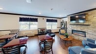 Located at 731 E Terra Cotta Ave in Crystal Lake, Illinois, this single-floor communal living room offers a well-appointed and inviting gathering space. The room is anchored by a striking stacked-stone fireplace with a sleek linear electric insert and a wall-mounted flat-screen television above, serving as the natural focal point of the space. Rich hardwood flooring runs throughout, complemented by crisp white walls accented with bold dark crown molding and a contrasting chair rail, lending the room a polished and cohesive aesthetic. The layout is thoughtfully divided into distinct functional zones. A cozy lounge area near the fireplace features a cluster of upholstered blue armchairs arranged around a round wooden coffee table, creating an intimate setting for conversation or relaxation. Open shelving units on either side of the fireplace add decorative storage and visual balance. A separate dining and casual seating area occupies the opposite side of the room, furnished with dark wood bistro-style tables and black chairs, ideal for group activities or casual dining. Multiple large windows dressed with dark curtains allow natural light to fill the space while offering views of the surrounding outdoor environment. Recessed lighting provides warm, even illumination throughout. The overall ambiance is comfortable, functional, and welcoming.