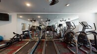 This single-floor fitness center is thoughtfully designed to accommodate a wide range of workout needs. The cardio zone is anchored by a row of Matrix treadmills and elliptical trainers, all positioned in front of expansive floor-to-ceiling mirrors that visually amplify the space and allow users to monitor their form. Dual-blade ceiling fans are distributed throughout, providing consistent airflow and comfort during workouts, complemented by recessed lighting that keeps the environment bright and energetic. The strength training area offers a selection of resistance machines, a multi-use weight bench, a power rack, and free-weight accessories, giving users a comprehensive suite of equipment for full-body conditioning. Exercise balls in multiple sizes are also available, supporting flexibility and functional fitness routines. A wall-mounted flat-screen television adds an entertainment element to the cardio section. The flooring throughout the facility consists of a bold red and gray patterned carpet tile, lending the space a modern, dynamic aesthetic. Structural columns are integrated into the open layout, and interior windows allow for natural light to filter through from adjacent areas. The clean white walls and well-organized equipment arrangement contribute to a welcoming and functional fitness environment.