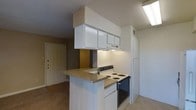 This single-floor unit centers around a compact, functional kitchen that flows openly into the adjacent living and entry areas. The kitchen is anchored by a peninsula with speckled laminate countertops, offering casual seating or prep space. White upper cabinetry with a built-in range hood overhead maximizes vertical storage, while a freestanding electric coil range sits neatly at the end of the peninsula. A full-height white refrigerator is tucked alongside the cabinetry, completing the kitchen's essentials. Fluorescent overhead lighting brightens the workspace efficiently. The kitchen opens toward the entryway, where a wood-panel front door with a standard deadbolt is visible, contributing to the unit's straightforward layout. The flooring transitions between the kitchen and adjacent living area, with light-toned carpet visible in the surrounding spaces. The walls feature a warm neutral palette, lending a modest, understated ambiance throughout. The single-floor layout ensures easy navigation, with the open sightlines between the kitchen and entry creating a sense of connectedness within the space.