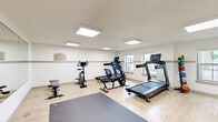 Located at 2401 6th St in Lake Charles, Louisiana, this fitness center is a well-appointed, single-room exercise facility designed to support a variety of workout routines. The space features light wood laminate flooring throughout, complemented by crisp white walls accented with a neutral mid-wall stripe, giving the room a clean and modern aesthetic.

Overhead, a series of flush-mounted LED panel lights provide bright, even illumination across the entire floor, while multiple windows along the walls allow natural light to filter in, creating an open and energizing atmosphere. A large wall-to-wall mirror along one side of the room visually expands the space and allows users to monitor their form during workouts.

The fitness center is equipped with a well-rounded selection of cardio machines, including treadmills, a stationary bike, a rowing-style machine, and an Endurance E5000 elliptical cross-trainer. A medicine ball rack with assorted weighted balls adds versatility for functional training, and a large exercise mat on the floor provides a dedicated area for stretching or floor-based workouts. A weight bench is also present for strength training.

Fitness center rules are posted on the wall, and seating is available for rest between sets. The room is accessible via a standard door, with an emergency exit sign clearly visible, ensuring safety compliance. Overall, the space is thoughtfully organized to maximize utility within its compact footprint.