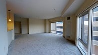 This single-floor space presents a clean, open layout centered around a generously sized living area finished with soft, light-toned wall-to-wall carpeting. The room is bathed in natural light, drawn in through a large sliding glass door fitted with vertical blinds, which opens to a balcony overlooking an exterior view of surrounding structures. The walls are finished in a neutral cream tone, contributing to a bright and airy atmosphere throughout. Structural beam details are visible along the ceiling, adding subtle architectural character to the otherwise streamlined interior. A utility panel is mounted along one wall, alongside visible HVAC vents, indicating integrated climate control throughout the space. The open floor plan allows for versatile furniture arrangement and a sense of spaciousness. A secondary doorway visible toward the rear of the space suggests additional rooms or storage beyond the main living area. Overall, the space offers a functional and understated design, well-suited for a variety of uses, with its combination of natural light, neutral finishes, and practical amenities creating a comfortable and adaptable environment.