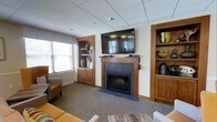 This inviting community room is thoughtfully designed to foster comfort and connection. At the heart of the space is a tile-surround fireplace, which serves as a natural focal point and adds warmth to the room's overall ambiance. A large wall-mounted flat-screen television is positioned above the fireplace, making it an ideal spot for shared viewing and entertainment. Flanking the fireplace are two built-in wooden bookcases with cabinetry below, offering both display space and storage. The warm tones of the wood millwork complement the neutral walls and carpeted flooring, creating a cohesive and welcoming aesthetic. Comfortable upholstered seating, including accent chairs and a sofa, is arranged to encourage conversation and relaxation. Natural light filters in through windows dressed with horizontal blinds, brightening the space without sacrificing privacy. A drop ceiling with recessed lighting provides even illumination throughout. The room strikes a balance between a residential feel and a functional communal setting, making it a versatile space well-suited for social gatherings, casual lounging, or organized community events.