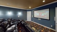 Located at 11655 Briar Forest Dr in Houston, Texas, this dedicated home theater room delivers an immersive cinematic experience within a thoughtfully designed single-floor space. The room is dressed in deep slate-blue walls that set a dramatic, theater-appropriate ambiance, complemented by warm gold-toned ceiling paint and crisp white crown molding that frames the space with a polished finish. Recessed ceiling lights and wall-mounted sconces provide adjustable ambient lighting, allowing for a true movie-going atmosphere. The focal point of the room is a large wall-mounted projection screen paired with a ceiling-mounted projector, creating a commanding viewing experience. Tiered rows of black leather recliner seats with built-in cup holders are arranged for optimal sightlines, comfortably accommodating multiple guests across both rows. In-wall speakers are positioned throughout the room to deliver full surround sound. A vibrant, decorative patterned carpet anchors the space and adds a playful cinematic flair underfoot. A standout feature near the entrance is a classic-style popcorn machine cart, reinforcing the authentic theater experience. An accessible restroom is conveniently located just off the entrance, making this space well-suited for extended private screenings and entertainment gatherings.