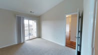 This single-floor space is characterized by its vaulted ceilings, which lend an airy, open quality to the interior. The main room is finished with neutral wall-to-wall carpeting and painted in a clean, light beige tone that enhances the sense of brightness throughout. A sliding glass door dominates one wall, providing direct access to a private outdoor balcony and allowing natural light to fill the space. The balcony features a classic railing and overlooks an exterior view with surrounding structures and trees visible in the distance. Ceiling-mounted HVAC vents indicate a central air system integrated into the vaulted ceiling design. An adjacent room, visible through a doorway, showcases warm hardwood flooring and continues the light, neutral color palette established throughout the space. The transition between the carpeted main room and the hardwood-floored adjoining room adds visual contrast and variety to the overall layout. Clean white trim and baseboards frame each room, contributing to a polished, well-maintained aesthetic. The overall ambiance is bright, functional, and move-in ready.