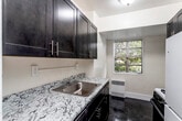This single-floor residence offers a well-organized layout designed for comfortable everyday living. The galley-style kitchen is a functional highlight, featuring rich dark espresso cabinetry with brushed nickel hardware, granite-patterned countertops, a stainless steel sink, and dark terrazzo-style flooring. A window at the end of the kitchen allows natural light to filter in, with views of surrounding greenery. The kitchen is equipped with a gas range and a full-size refrigerator, along with ample upper and lower cabinet storage.

The living areas are finished with neutral wall-to-wall carpeting and warm cream-toned walls, creating a bright and welcoming atmosphere. The spacious main living area connects to multiple rooms, offering a flexible and open feel. The bedrooms are generously sized, each featuring carpeted floors, windows that bring in natural light, and closet space for storage. Baseboard heating units are present throughout the residence.

The bathroom is finished with classic white subway tile surrounding a full soaking tub with an overhead shower, complemented by terrazzo-style flooring and clean, neutral walls. Overall, the space presents a practical and cohesive design with consistent finishes throughout.