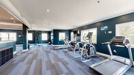 Located at 6320 Logans Ln in Lynchburg, Virginia, this well-appointed fitness center offers a generously sized, single-floor layout designed to accommodate a comprehensive range of workout activities. The interior is finished with a bold deep navy and teal color palette across the walls, complemented by clean white crown molding and a bright white ceiling with recessed lighting that keeps the space feeling open and energizing. Durable, patterned carpet tiles in blue and grey tones cover the floors throughout, providing both comfort and practicality underfoot. Multiple windows line the walls, inviting abundant natural light and offering views of the surrounding exterior. The cardio zone is equipped with treadmills, elliptical trainers, and cycling bikes, while a dedicated strength training area features a multi-station cable machine, weight benches, and a full rack of kettlebells. Large wall mirrors in the strength section enhance the sense of space and allow for proper form monitoring. A stretching and functional fitness area with yoga mats rounds out the facility. Exposed ductwork in the strength zone adds an industrial touch, while a decorative stone accent column serves as a distinctive architectural focal point at the heart of the space.