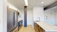 This single-floor space masterfully combines historic industrial character with contemporary design elements. Exposed red brick walls serve as a striking architectural focal point throughout, contrasting beautifully with crisp white walls and light hardwood-style flooring that runs continuously through the unit.

The kitchen is thoughtfully designed in a galley layout, featuring two-tone cabinetry pairing white upper cabinets with warm wood-toned lower cabinets and black hardware. Stainless steel appliances include a French door refrigerator, range with oven, and microwave. A white quartz countertop extends along one side, housing a stainless steel sink with a modern arc faucet. White subway tile backsplash adds a clean, classic detail to the cooking area. A distinctive dark structural column stands as a notable architectural element within the kitchen space.

The living and bedroom areas showcase original exposed brick walls and large windows that bring in natural light while offering urban views. Exposed ductwork along the ceiling reinforces the industrial aesthetic.

The bathroom features large-format gray tile flooring, floor-to-ceiling tiled walls, and a frameless glass-enclosed walk-in shower, delivering a clean and modern finish that complements the overall design of the space.