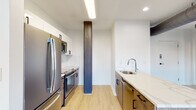 This single-floor space masterfully combines historic industrial character with contemporary design elements. Exposed red brick walls serve as a striking architectural focal point throughout, contrasting beautifully with crisp white walls and light hardwood-style flooring that runs continuously through the unit.

The kitchen is thoughtfully designed in a galley layout, featuring two-tone cabinetry pairing white upper cabinets with warm wood-toned lower cabinets and black hardware. Stainless steel appliances include a French door refrigerator, range with oven, and microwave. A white quartz countertop extends along one side, housing a stainless steel sink with a modern arc faucet. White subway tile backsplash adds a clean, classic detail to the cooking area. A distinctive dark structural column stands as a notable architectural element within the kitchen space.

The living and bedroom areas showcase original exposed brick walls and large windows that bring in natural light while offering urban views. Exposed ductwork along the ceiling reinforces the industrial aesthetic.

The bathroom features large-format gray tile flooring, floor-to-ceiling tiled walls, and a frameless glass-enclosed walk-in shower, delivering a clean and modern finish that complements the overall design of the space.