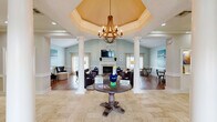 Located at 7900 Berryhill Place in Montgomery, Alabama, this single-floor space offers a thoughtfully designed interior that balances elegance with comfort. The hallway serves as a grand entry point, showcasing travertine tile flooring, tall white columns, and a tray ceiling adorned with a rustic wood-and-iron chandelier. Large glass panel partitions allow sightlines into adjoining areas, creating an open and connected feel throughout.

The spacious living room is a standout feature, boasting soaring vaulted ceilings that amplify the sense of openness. A classic white-surround fireplace serves as the room's focal point, complemented by hardwood flooring and soft, neutral wall tones in a calming sage-white palette. Large windows dressed with sheer linen curtains invite abundant natural light, while a ceiling fan adds practical comfort. The room accommodates generous seating arrangements, making it ideal for gathering and relaxation.

A cozy sitting area with warm hardwood floors features wainscoting and soft blue-grey walls, providing an intimate retreat within the larger space. Throughout, the design reflects a cohesive coastal-inspired aesthetic with carefully chosen artwork and understated décor. The overall ambiance is refined yet inviting, making this a comfortable and functional communal environment.