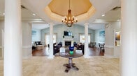 Located at 7900 Berryhill Place in Montgomery, Alabama, this single-floor space offers a thoughtfully designed interior that balances elegance with comfort. The hallway serves as a grand entry point, showcasing travertine tile flooring, tall white columns, and a tray ceiling adorned with a rustic wood-and-iron chandelier. Large glass panel partitions allow sightlines into adjoining areas, creating an open and connected feel throughout.

The spacious living room is a standout feature, boasting soaring vaulted ceilings that amplify the sense of openness. A classic white-surround fireplace serves as the room's focal point, complemented by hardwood flooring and soft, neutral wall tones in a calming sage-white palette. Large windows dressed with sheer linen curtains invite abundant natural light, while a ceiling fan adds practical comfort. The room accommodates generous seating arrangements, making it ideal for gathering and relaxation.

A cozy sitting area with warm hardwood floors features wainscoting and soft blue-grey walls, providing an intimate retreat within the larger space. Throughout, the design reflects a cohesive coastal-inspired aesthetic with carefully chosen artwork and understated décor. The overall ambiance is refined yet inviting, making this a comfortable and functional communal environment.