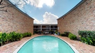 This property is centered around a striking outdoor courtyard pool, serving as the focal point of the space. The pool features a curved design with a brick-paved surround, complemented by lounge seating and decorative greenery that frames the area with a welcoming, resort-like atmosphere. The surrounding brick walls rise on either side of the courtyard, creating a sense of enclosure and privacy while opening to the sky above, allowing for abundant natural light throughout the day. A multi-level building serves as the backdrop, featuring covered walkways and large windows that overlook the pool area. The architectural design is classic and durable, with warm-toned brick construction throughout that lends a timeless character to the overall aesthetic. Mature shrubs and plantings line the perimeter, softening the hardscape and adding natural texture to the courtyard. The combination of open sky, brick architecture, and manicured landscaping creates a cohesive and inviting outdoor environment well-suited for relaxation and leisure.