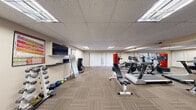 This single-room fitness space offers a fully equipped workout environment within a compact, efficiently utilized footprint. The room features a drop-panel ceiling with recessed fluorescent lighting that evenly illuminates the entire area, complemented by vertical blind-covered sliding doors along one wall that allow natural light to filter in. Striped, low-pile carpet tiles cover the floor throughout, providing a durable and practical surface suited to fitness use.

The space is organized to accommodate a range of exercise modalities. Cardio equipment, including treadmills and stationary bikes, is positioned along one side of the room. A multi-station cable and weight machine occupies another corner, alongside a flat adjustable workout bench. Free weights are well-represented, with both a horizontal dumbbell rack and a vertical tower rack holding a graduated set of dumbbells, as well as medicine balls arranged along the baseboard. A wall-mounted flat-screen television is positioned above a small media shelf, with instructional fitness posters flanking it. Sanitation dispensers are mounted nearby for user convenience. Large wall mirrors on two sides visually expand the space and assist with form monitoring. The overall ambiance is clean, functional, and purpose-driven.