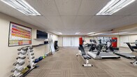 This single-room fitness space offers a fully equipped workout environment within a compact, efficiently utilized footprint. The room features a drop-panel ceiling with recessed fluorescent lighting that evenly illuminates the entire area, complemented by vertical blind-covered sliding doors along one wall that allow natural light to filter in. Striped, low-pile carpet tiles cover the floor throughout, providing a durable and practical surface suited to fitness use.

The space is organized to accommodate a range of exercise modalities. Cardio equipment, including treadmills and stationary bikes, is positioned along one side of the room. A multi-station cable and weight machine occupies another corner, alongside a flat adjustable workout bench. Free weights are well-represented, with both a horizontal dumbbell rack and a vertical tower rack holding a graduated set of dumbbells, as well as medicine balls arranged along the baseboard. A wall-mounted flat-screen television is positioned above a small media shelf, with instructional fitness posters flanking it. Sanitation dispensers are mounted nearby for user convenience. Large wall mirrors on two sides visually expand the space and assist with form monitoring. The overall ambiance is clean, functional, and purpose-driven.