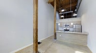 This single-floor loft seamlessly blends industrial heritage with contemporary design, creating a space that is both characterful and functional. Soaring ceilings with exposed wooden beams and a warm timber ceiling define the open layout, lending an expansive, airy quality to the interior. Original structural columns in reclaimed wood anchor the space, serving as striking architectural focal points that nod to the building's storied past.

Polished concrete floors run throughout, providing a sleek, durable surface that complements the raw industrial aesthetic while remaining easy to maintain. The open-plan living area flows naturally into a well-appointed kitchen, where light wood cabinetry pairs elegantly with stainless steel appliances, including a range and an over-the-range microwave. A generous kitchen island with a stone-look countertop offers ample prep space and doubles as a casual dining or entertaining surface.

Pendant lighting and industrial-style hanging fixtures illuminate the space thoughtfully, highlighting the interplay between the rustic structural elements and the clean, modern finishes. The neutral grey walls provide a versatile backdrop, making the space highly adaptable to a variety of interior styles. Overall, this loft offers a distinctive atmosphere that celebrates its architectural roots while delivering modern comfort and utility.