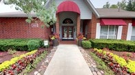This single-story brick building presents a polished and inviting exterior that immediately conveys professionalism and attention to detail. The entrance is highlighted by a bold red arched awning framing a set of elegant double doors topped with a decorative arched transom window, allowing natural light to filter into the entryway. A well-maintained concrete walkway leads visitors to the front door, flanked by meticulously curated landscaping that includes vibrant seasonal flower arrangements in large planters, neatly trimmed hedges, and decorative river rock borders that add texture and visual interest. Colorful blooms in shades of red, yellow, and orange provide a warm, cheerful atmosphere that enhances the building's curb appeal. The brick facade is complemented by light gray siding accents and white-trimmed windows, some of which feature matching red awnings that create a cohesive and attractive aesthetic. A classic wood bench near the entrance offers a welcoming touch for visitors. The surrounding mature trees provide natural shade and a lush green backdrop, contributing to the overall pleasant and approachable character of this well-appointed commercial space.