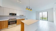 Located on the penthouse level at 999 NW Seventh St in Miami's vibrant urban core, this one-bedroom, one-bathroom residence offers a thoughtfully designed open floor plan with clean, modern finishes throughout. The open-concept kitchen serves as the heart of the home, showcasing a two-tone design with white upper cabinetry and warm wood lower cabinets, a white quartz countertop island, stainless steel appliances, a tiled backsplash, and a striking trio of industrial-style pendant lights overhead. The soaring ceilings amplify the sense of space, and large sliding glass doors lead to a private balcony with expansive views of Miami's skyline and lush tree canopy below. Light-toned large-format tile flooring flows throughout, brightening the interior. The bedroom is generously sized, benefiting from floor-to-ceiling windows that frame sweeping outdoor views and bathe the room in natural light. Adjacent to the bedroom are two separate doors — one leading to a spacious walk-in closet with built-in wire shelving, and another into the bathroom. The bathroom features a long wood-toned vanity with a white quartz countertop, a double sink, a framed mirror, and access to a separate water closet with tile-clad walls. The overall ambiance is bright, airy, and contemporary.