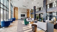 This contemporary amenity lounge is defined by its dramatic double-height ceilings and expansive floor-to-ceiling windows that flood the interior with natural light and provide unobstructed views of the surrounding outdoor courtyard, complete with seating areas and vibrant umbrellas. The open-plan layout is anchored by a striking mixed-material flooring design, combining light hardwood in a herringbone pattern with bold geometric inlays that guide movement through the space.

Architectural interest is provided by prominent textured stone-clad columns, a large-scale reclaimed wood accent wall, and elegant marble wall panels that add warmth and depth. Pendant lighting clusters descend from the soaring ceiling, creating intimate pockets of light throughout the lounge. A mezzanine level, accessible via an industrial-style staircase with metal railings, overlooks the main floor and adds a sense of vertical grandeur.

The lounge is furnished with a curated mix of modern seating — including plush sofas, bold emerald green and cobalt blue accent chairs, and sculptural side tables — arranged to encourage both social gathering and quiet retreat. A wall-mounted flat-screen TV and a billiards table provide entertainment options, while a dedicated seating nook near the window wall offers a more intimate setting. Glazed doors open directly to the outdoor courtyard, seamlessly connecting the interior and exterior amenity spaces.