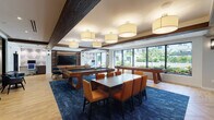 Located at 1365–1367 S Dixie Hwy in Coral Gables, Florida, this single-floor amenity lounge is a thoughtfully designed social and recreational space that blends modern sophistication with inviting warmth. The open-plan layout is anchored by rich herringbone wood flooring and bold exposed wooden ceiling beams that add a striking architectural contrast to the bright white ceiling above.

The game area serves as a central focal point, featuring a full-size billiards table and a shuffleboard table, both crafted in warm wood tones and set atop a deep navy patterned area rug. A large dining table surrounded by upholstered orange chairs provides ample space for group gatherings, while a counter-height bar with woven-seat stools offers a more casual seating option.

The lounge area is outfitted with a leather sofa, accent chairs in blue and cognac, and a round brass coffee table, all oriented toward a large wall-mounted flat-screen TV set within a custom built-in wood entertainment unit with integrated LED accent lighting.

A well-appointed kitchen features a broad white quartz island countertop with bar seating, dark wood cabinetry, a mosaic tile backsplash, and stainless steel appliances. Oversized windows throughout flood the space with natural light and offer views of lush tropical landscaping, creating a seamless connection to the South Florida outdoor environment.