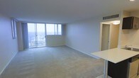 This single-floor space offers an open, airy layout filled with abundant natural light. The main living area is defined by its clean, minimalist design, featuring smooth white walls, light-toned carpeted flooring, and a white flat ceiling with integrated ventilation. A standout feature is the expansive floor-to-ceiling sliding glass door system along one wall, which floods the interior with daylight and provides direct access to a balcony. From this elevated vantage point, the views extend across a cityscape and out toward the ocean horizon, lending the space a striking sense of openness and connection to the surrounding environment. A partial countertop visible at the edge of the living area suggests an adjacent kitchen or dining space, hinting at a functional and practical layout. A doorway on the opposite side opens to additional rooms beyond, indicating a well-organized floor plan. The overall ambiance is calm and understated, with neutral tones throughout creating a versatile backdrop suited to a variety of uses. The combination of generous natural light, elevated views, and a straightforward, open layout makes this a bright and inviting space.