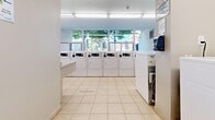 Located at 2797 Battleford Rd in Mississauga, Ontario, this shared laundry facility offers a well-organized and functional space designed to serve building residents efficiently. The room is finished with light-toned ceramic tile flooring throughout, and bright fluorescent lighting ensures clear visibility across the entire area.

The main laundry zone is equipped with a generous lineup of front-loading washing machines and top-loading dryers, arranged in rows to maximize capacity. Large windows along one wall allow natural light to filter in, offering views of the surrounding greenery and contributing to a clean, open atmosphere. A built-in folding counter with an integrated utility sink provides a convenient workspace for handling laundry, while a paper towel dispenser is mounted nearby for added practicality.

A separate alcove within the space is dedicated to household hazardous waste and electronics recycling, featuring clearly labeled drop-off bins and posted safety guidelines — reflecting a commitment to environmentally responsible practices. Wall-mounted informational signage throughout the facility provides instructions for machine operation and recycling procedures.

The layout is thoughtfully arranged into distinct functional zones, making efficient use of the available floor area. Overall, the facility presents a clean, bright, and well-maintained environment that prioritizes convenience and practicality for everyday use.