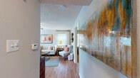 This single-story residence in Palm Harbor, Florida offers a thoughtfully designed layout with distinct living spaces connected by wood-look flooring throughout the main areas. The living room serves as the heart of the home, featuring a textured ceiling, natural light from a window dressed with patterned curtains, and wall space well-suited for artwork and decorative accents. A hallway adorned with a large statement painting leads seamlessly from the entry into the main living area.

The dining area benefits from generous window coverage, providing an open, airy feel with views of the surrounding greenery. Track lighting overhead and an adjacent built-in bar ledge add functional character to the space. The galley-style kitchen is equipped with light wood cabinetry, dark countertops, black appliances including a range and refrigerator, and access to a dedicated laundry area with an in-unit washer.

Both bedrooms feature plush carpet, ceiling fans, and ample natural light. One bedroom showcases a bold teal accent wall and a reach-in closet, while the second offers a calmer, neutral palette. The bathroom includes a full tub-shower combination with clean white tile finishes.