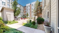 Located at 3001 Fox St in Denver, Colorado, this outdoor courtyard is a well-appointed communal space nestled within a multi-story residential complex. The courtyard is defined by a broad, smooth concrete plaza that serves as the central gathering area, bordered by thoughtfully designed landscaping that includes mature trees, ornamental shrubs, flowering plants, and decorative rock beds — all contributing to a lively yet low-maintenance aesthetic well-suited to the Colorado climate.

Multiple seating zones are distributed throughout the space to accommodate a variety of uses. Sleek chaise lounges invite residents to relax and enjoy the abundant Colorado sunshine, while a pergola-covered dining area provides shaded outdoor seating ideal for casual meals or social gatherings. Vibrant red Adirondack chairs add a bold pop of color and a welcoming, relaxed atmosphere. Concrete benches and additional potted plantings further enrich the space.

The surrounding building facades — clad in a combination of corrugated metal panels and warm wood-toned siding in grey and rust hues — frame the courtyard on multiple sides, creating a sheltered and cohesive environment. A small utility structure is integrated discreetly within the landscape. The open-sky exposure ensures excellent natural light throughout the day, making this courtyard a functional and attractive outdoor amenity.