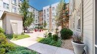 Located at 3001 Fox St in Denver, Colorado, this outdoor courtyard is a well-appointed communal space nestled within a multi-story residential complex. The courtyard is defined by a broad, smooth concrete plaza that serves as the central gathering area, bordered by thoughtfully designed landscaping that includes mature trees, ornamental shrubs, flowering plants, and decorative rock beds — all contributing to a lively yet low-maintenance aesthetic well-suited to the Colorado climate.

Multiple seating zones are distributed throughout the space to accommodate a variety of uses. Sleek chaise lounges invite residents to relax and enjoy the abundant Colorado sunshine, while a pergola-covered dining area provides shaded outdoor seating ideal for casual meals or social gatherings. Vibrant red Adirondack chairs add a bold pop of color and a welcoming, relaxed atmosphere. Concrete benches and additional potted plantings further enrich the space.

The surrounding building facades — clad in a combination of corrugated metal panels and warm wood-toned siding in grey and rust hues — frame the courtyard on multiple sides, creating a sheltered and cohesive environment. A small utility structure is integrated discreetly within the landscape. The open-sky exposure ensures excellent natural light throughout the day, making this courtyard a functional and attractive outdoor amenity.