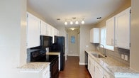 This 2-bedroom, 2-bathroom residence offers a thoughtfully designed single-floor layout that balances style and functionality. The kitchen is a standout feature, showcasing crisp white cabinetry that extends to the ceiling, granite-look countertops, and a classic subway tile backsplash. Black appliances — including a range, built-in microwave, refrigerator, and dishwasher — provide a bold contrast against the bright cabinetry. A stainless steel sink is positioned beneath a window, allowing natural light to fill the space. Track lighting overhead ensures the kitchen is well-lit throughout the day and evening. Rich hardwood-style flooring runs through the kitchen and into the hallway, adding warmth and a cohesive flow throughout the home. The hallway connects the main living areas to the bedrooms and bathrooms, providing a practical and open corridor. A textured ceiling adds a subtle architectural detail throughout the space. Overall, the home presents a clean, move-in-ready aesthetic with durable finishes, ample storage, and a layout that makes efficient use of every square foot.