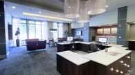 This sophisticated single-floor commercial space makes a strong first impression with its thoughtfully designed interior. At the heart of the space is a polished reception and workspace area outfitted with sleek, dark-wood workstations topped with crisp white surfaces, each equipped with computer monitors for efficient service. Dramatic cascading chandelier pendants hang from a coffered ceiling, adding a refined, upscale ambiance to the environment.

The space is bathed in abundant natural light, courtesy of expansive floor-to-ceiling windows dressed with sheer white curtains that soften the incoming daylight. A comfortable lounge area sits adjacent to the main workspace, furnished with deep-toned armchairs and small tables, providing a welcoming spot for visitors. The flooring throughout features slate-toned tile, lending a grounded, contemporary feel that complements the overall color palette of warm grays, rich browns, and soft whites.

Wall-mounted flat-screen displays and carefully selected artwork punctuate the walls, contributing both functionality and visual interest. Recessed lighting supplements the natural light, ensuring a bright and professional atmosphere throughout. The overall design balances modern elegance with practical functionality, creating a welcoming and highly professional environment.