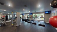 This single-floor fitness center is thoughtfully designed to support a wide range of workout routines. The space features durable dark rubber flooring throughout, providing both safety and functionality across all exercise zones. A row of treadmills and elliptical trainers lines the windowed wall, where large windows invite natural light and offer views of lush outdoor greenery, creating an energizing atmosphere during cardio sessions. Wall-mounted flat-screen televisions are positioned above the cardio equipment, keeping users entertained throughout their workouts. A full-length mirrored wall extends across one side of the room, visually expanding the space and allowing users to monitor their form during exercise. The strength training area includes a multi-station cable and weight machine, a rack of dumbbells in varying weights, and a medicine ball storage rack stocked with balls of multiple sizes for functional training. Additional stability and exercise balls are available for versatile conditioning. The facility is accessed through elegant French doors, and a water fountain station is conveniently located near the entrance. Recessed lighting illuminates the entire space, complementing the natural light from the windows. The neutral gray walls and textured ceiling contribute to a clean, modern aesthetic throughout this well-appointed fitness facility.