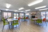 This single-floor community space in Phenix City, Alabama, is designed to accommodate gatherings and social interaction with a bright, welcoming atmosphere. The open main room is expansive and well-lit, featuring light wood-look flooring throughout that lends warmth and cohesion to the interior. A striking whitewashed brick fireplace serves as a central focal point, complete with a wood mantel, decorative accessories, and a classic fireplace screen, adding character and charm to the space.

Multiple round and rectangular white tables are paired with vibrant green and orange chairs, creating a lively, energetic aesthetic that encourages collaboration and conversation. Flush-mounted and track lighting fixtures illuminate the space evenly, while large windows fitted with horizontal blinds invite ample natural light and offer views of the outdoor surroundings, including what appears to be a pool area.

The kitchenette is thoughtfully appointed with white shaker-style cabinetry, granite countertops, a white subway tile backsplash, and stainless steel appliances. Framed artwork displayed along the neutral-toned walls adds a polished, finished look. Multiple doorways provide convenient access throughout, making this a functional and inviting communal environment.