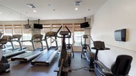 This fitness center offers a comprehensive workout environment designed to support a wide range of exercise routines. The space is bright and welcoming, with multiple windows dressed in woven bamboo shades that allow natural light to filter in while maintaining a comfortable atmosphere. Recessed lighting throughout ensures the room is well-lit at all times.

The cardio zone is prominently featured and includes multiple treadmills, elliptical trainers, and stationary bikes — each positioned to face wall-mounted flat-screen televisions for an engaging workout experience. The machines are modern and well-maintained, reflecting a high standard of facility upkeep.

A dedicated free weights area provides a tiered dumbbell rack stocked with a full range of weights, accompanied by adjustable workout benches suitable for a variety of strength training exercises. A multi-functional cable machine anchors the strength training section, offering versatility for users of all fitness levels.

Floor-to-ceiling mirrors line one side of the room, visually expanding the space and allowing users to monitor their form during workouts. The durable rubber flooring provides both safety and comfort underfoot. The overall design is clean, functional, and purposefully laid out to maximize usability while maintaining an open, uncluttered feel throughout.