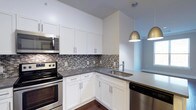 Unit 405 at 333 Central Avenue offers a well-appointed single-floor layout designed with modern finishes and thoughtful details throughout. The kitchen serves as a standout feature, showcasing crisp white shaker-style cabinetry that extends to the ceiling, maximizing storage space. A mosaic tile backsplash in neutral tones of gray and white adds texture and visual interest, complementing the sleek dark countertops that run along the L-shaped workspace. Stainless steel appliances — including a range, built-in microwave, and dishwasher — lend a polished, contemporary feel to the cooking space. A undermount sink with a modern faucet is positioned at the peninsula, which opens toward the adjacent living area, creating an open and connected layout ideal for entertaining. Stylish metallic dome pendant lights hang above the peninsula, casting warm, focused light and adding a refined design touch. The rich dark hardwood flooring contrasts beautifully with the bright white cabinetry, grounding the space with warmth and character. Overall, the unit presents a clean, contemporary aesthetic with high-quality finishes that balance both functionality and style.