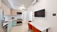 Located at 360 N Hayworth Ave in Los Angeles' desirable Mid-Wilshire neighborhood, this modern single-floor unit offers five bedrooms and three bathrooms with a clean, minimalist aesthetic throughout. Light hardwood flooring runs continuously across the entire space, complementing the crisp white walls and creating a bright, cohesive feel.

The combined kitchen and living area serves as the central hub of the unit. The kitchen features light wood cabinetry with sage green lower cabinets, a stainless steel gas range with an overhead microwave, and recessed lighting. A sleek white dining table with vibrant orange chairs anchors the living area, alongside a wall-mounted TV and a ductless mini-split climate control system.

A clean hallway connects the common areas to the five bedrooms, each furnished with a platform bed featuring a warm wood frame and individual closets for storage. Several bedrooms benefit from natural light through large windows offering leafy outdoor views. The primary bedroom is notably spacious and includes a dedicated closet area with built-in shelving.

The three bathrooms maintain the minimalist theme, showcasing textured wave-patterned wall tiles, large-format floor tiles, floating wood vanities with white countertops, frameless glass shower enclosures, and matte black fixtures throughout.