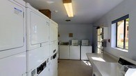 This shared laundry facility is designed with practicality and efficiency in mind, providing all the essentials for a convenient laundry experience. The space is outfitted with multiple top-loading washing machines lined along the back wall, complemented by a set of stacked dryers positioned along the side wall, offering generous overall capacity for users. A folding counter runs along one side, providing a dedicated surface for sorting and folding clothes. The room is finished with light gray walls and a neutral floor, lending a clean and straightforward aesthetic throughout. Overhead fluorescent lighting ensures the space is brightly and evenly illuminated. A window along one wall allows natural light to filter in, adding a sense of openness to the room. The layout is thoughtfully organized, with clear pathways between machines to allow for easy access and comfortable use. A HVAC vent in the ceiling supports proper air circulation, keeping the environment fresh. Overall, this laundry facility is a well-maintained, no-frills space that prioritizes function and accessibility, making it a practical amenity for shared use.