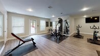 Located at 100 Marsh Point Dr in Hilton Head, SC, this single-floor fitness center offers a comprehensive and thoughtfully organized workout facility. The space is divided into two connected workout areas, both finished with durable light wood-look flooring and protective rubber mats beneath the equipment.

The main strength and cardio area features a multi-station cable machine flanked by a full wall of floor-to-ceiling mirrors, which amplify the sense of space while allowing users to monitor their form. This zone also includes a treadmill, elliptical trainer, rowing machine, adjustable weight bench, and a rack of free weights. Natural light filters in through multiple windows fitted with white shutters, and ceiling fans provide comfortable air circulation throughout.

An adjacent studio-style room is dedicated to cycling and group fitness, housing multiple spin bikes and additional cardio equipment including a stationary bike. A wall-mounted flat-screen television enhances the workout experience, while exercise balls and accessories stored in woven baskets add functional versatility for stretching and core training.

Both areas feature recessed lighting and a clean, neutral color palette — with one wall accented in a deep slate blue — creating a focused and energizing environment. The facility is accessible via an exterior door, making it convenient for community use.