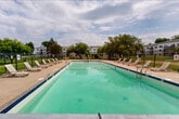 This outdoor community space is centered around a generously sized rectangular swimming pool with clear, blue-green water, offering an inviting retreat for residents. The pool is accessed through a secure black wrought-iron gate, with posted signage indicating that a pool pass is required for entry — ensuring a private and controlled environment for authorized users.

Surrounding the pool, a broad concrete sun deck provides ample space for relaxation, lined with multiple lounge chairs positioned to take full advantage of open skies and natural light. Picnic tables are conveniently situated near the pool area, offering casual outdoor dining and gathering spots. Safety features, including pool entry railings and life-saving equipment, are visibly in place throughout the deck.

The grounds extend well beyond the pool enclosure, with expansive, well-kept green lawns framed by mature deciduous trees that provide natural shade and a sense of seclusion. Paved walkways meander through the landscaped grounds, connecting the pool area to the surrounding residential buildings, which are multi-story structures featuring balconies and classic architectural detailing. Additional picnic tables are scattered across the open lawn areas, further enhancing the space's functionality for outdoor gatherings. The overall ambiance is open, relaxed, and community-oriented, offering a serene outdoor environment ideal for recreation and leisure.