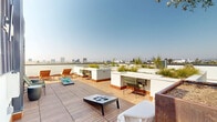 Located at 360 N Hayworth Ave in the heart of Los Angeles, this rooftop deck offers an impressive outdoor retreat with sweeping panoramic views of the city skyline, the Hollywood Hills, and the surrounding neighborhoods. The deck is finished with warm-toned hardwood tile flooring that extends across the entire space, lending a polished yet inviting aesthetic that complements the Southern California sunshine.

The layout is thoughtfully organized into distinct zones. A sun lounging area features multiple teak-style chaise lounges arranged for optimal sun exposure and city views. A casual social zone includes a curved built-in bench surrounding a central planter, creating a sheltered gathering spot with a fire feature and integrated greenery. Additional seating nooks with modern chairs and low tables are positioned throughout to take advantage of the varying vantage points.

Large white stucco planters are positioned around the perimeter and throughout the deck, filled with ornamental grasses, native shrubs, and a mature olive tree, adding lush, natural texture to the urban setting. A built-in counter station provides a functional outdoor workspace or bar area. Recreational options include cornhole game setups ready for use.

A dark metal mechanical enclosure anchors one section of the rooftop, providing a design contrast to the crisp white parapet walls. The open sky and unobstructed sightlines make this rooftop an exceptional amenity space.