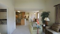 This single-story residence is designed around an open-concept floor plan that seamlessly integrates the main living spaces. The living area is carpeted and comfortably proportioned, anchored by a green upholstered armchair and accented with decorative plants and a floor lamp, creating a welcoming atmosphere. A ceiling fan overhead provides both function and a subtle design element.

The layout flows naturally into a dining area, which accommodates a full dining table and chairs beneath a matching ceiling fan. A window in this zone allows natural light to filter in, brightening the space. Framed wall art adds visual interest throughout.

The kitchen is partially open to the living area, separated by a breakfast bar with counter seating. It features wood-toned cabinetry, a bold patterned accent wall, and overhead lighting that illuminates the workspace. Appliances are integrated within the cabinetry layout. A hallway visible from the main living space suggests additional rooms beyond, contributing to the overall sense of a well-connected, functional floor plan with consistent neutral tones and warm finishes throughout.