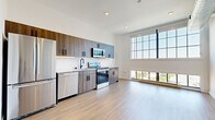 This thoughtfully designed single-floor unit offers a bright, open-plan layout that maximizes its compact footprint. The living area is defined by dramatic floor-to-ceiling grid windows that flood the space with natural light and frame expansive outdoor views of the surrounding cityscape. Exposed spiral ductwork adds an industrial loft character to the soaring ceilings, while light-toned hardwood flooring runs seamlessly throughout, enhancing the sense of openness and continuity.

The kitchen is fully equipped and efficiently arranged along a single wall, featuring wood-grain cabinetry with matte black hardware, a white subway tile backsplash, light stone countertops, and a full suite of stainless steel appliances including a refrigerator, electric range, microwave, and dishwasher. Recessed lighting overhead ensures the kitchen remains bright and functional.

The bathroom presents a clean, modern aesthetic with large-format tile flooring, a floating vanity with a white quartz countertop, an undermount sink with a matte black faucet, and a well-placed wall sconce that provides warm, even lighting. A mirror above the vanity enhances the sense of space. Throughout the unit, crisp white walls and minimalist finishes create a cohesive, contemporary ambiance suited to modern living.