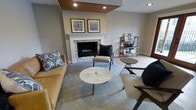 This single-floor living space showcases a carefully curated interior that blends contemporary and mid-century modern design elements. The focal point of the room is a striking fireplace framed by a classic white mantel and clad in stacked stone veneer, adding both texture and warmth to the space. Above the mantel, two coordinating framed artworks in blue and gray tones contribute a cohesive, polished aesthetic.

A distinctive wood-paneled ceiling detail with recessed lighting draws the eye upward, adding architectural interest and warmth to the otherwise neutral gray palette of the walls and trim. Light-toned hardwood flooring transitions seamlessly to a large area rug in muted blue-gray, anchoring the seating arrangement.

The furnishings include a rich caramel leather sofa, a pair of mid-century inspired accent chairs in taupe with dark frames, and a sculptural round marble-top coffee table. A wood ladder-style bookshelf along one wall provides functional display space. The overall ambiance is sophisticated yet approachable, with a neutral color scheme accented by warm wood tones and layered textures throughout.