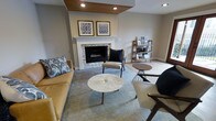 This single-floor living space showcases a carefully curated interior that blends contemporary and mid-century modern design elements. The focal point of the room is a striking fireplace framed by a classic white mantel and clad in stacked stone veneer, adding both texture and warmth to the space. Above the mantel, two coordinating framed artworks in blue and gray tones contribute a cohesive, polished aesthetic.

A distinctive wood-paneled ceiling detail with recessed lighting draws the eye upward, adding architectural interest and warmth to the otherwise neutral gray palette of the walls and trim. Light-toned hardwood flooring transitions seamlessly to a large area rug in muted blue-gray, anchoring the seating arrangement.

The furnishings include a rich caramel leather sofa, a pair of mid-century inspired accent chairs in taupe with dark frames, and a sculptural round marble-top coffee table. A wood ladder-style bookshelf along one wall provides functional display space. The overall ambiance is sophisticated yet approachable, with a neutral color scheme accented by warm wood tones and layered textures throughout.