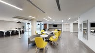 This thoughtfully designed single-floor communal space offers a bright, open-concept layout that seamlessly blends style with functionality. The interior is defined by clean white ceilings with recessed linear lighting that casts a consistent, professional glow throughout the room. Wide-plank wood-style flooring adds warmth and continuity across the space, complementing the neutral tones of the walls and structural columns.

The lounge area is furnished with a curated mix of modern seating, including curved gray sectional sofas, round white tables, and vibrant yellow accent chairs that inject energy and personality into the environment. Multiple seating clusters are arranged throughout the space, encouraging both casual conversation and collaborative gatherings.

Expansive floor-to-ceiling windows along one side of the space flood the interior with natural light and offer views of the surrounding urban landscape. A wall-mounted flat-screen display is centrally positioned, making the space suitable for presentations or shared viewing.

Additional high-top tables and bar seating are visible near the window line, providing versatile options for informal meetings or individual work. The overall ambiance is clean, contemporary, and welcoming — a well-appointed communal environment designed to support a variety of social and professional activities.